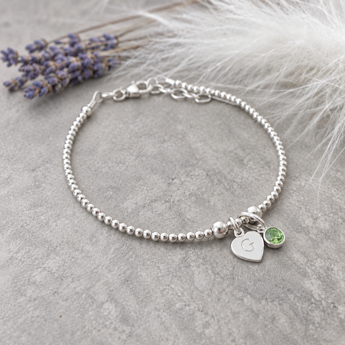 A silver-toned bracelet with a heart-shaped charm and a small gemstone is displayed on a gray surface, accompanied by a bunch of purple flowers.
