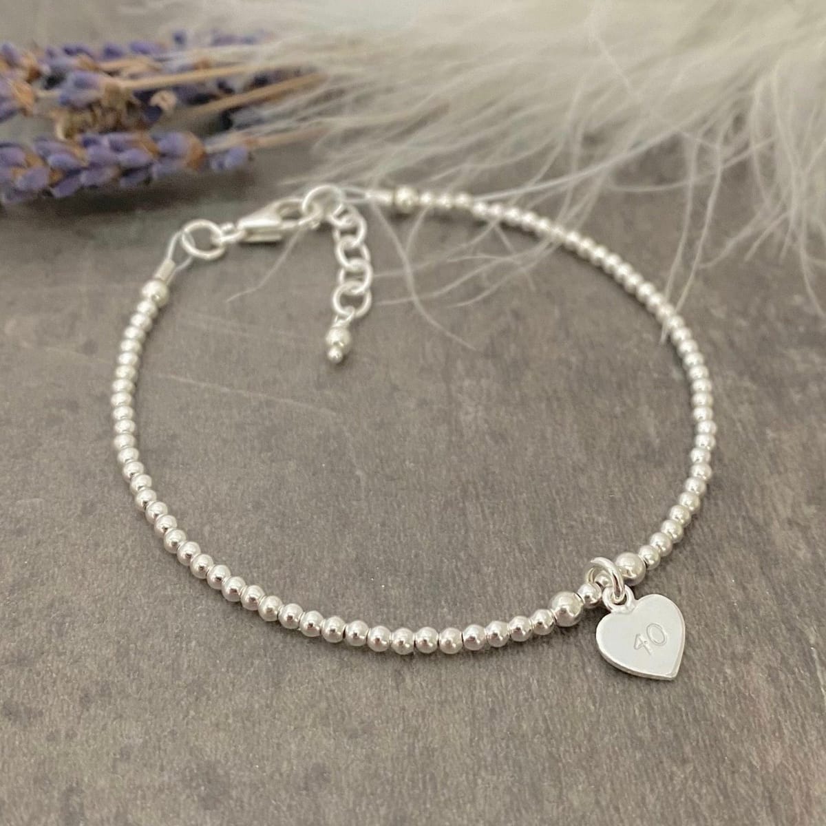 A silver bracelet with a heart-shaped charm and the number 10 engraved on it, placed on a gray surface with a feather-like object in the background.