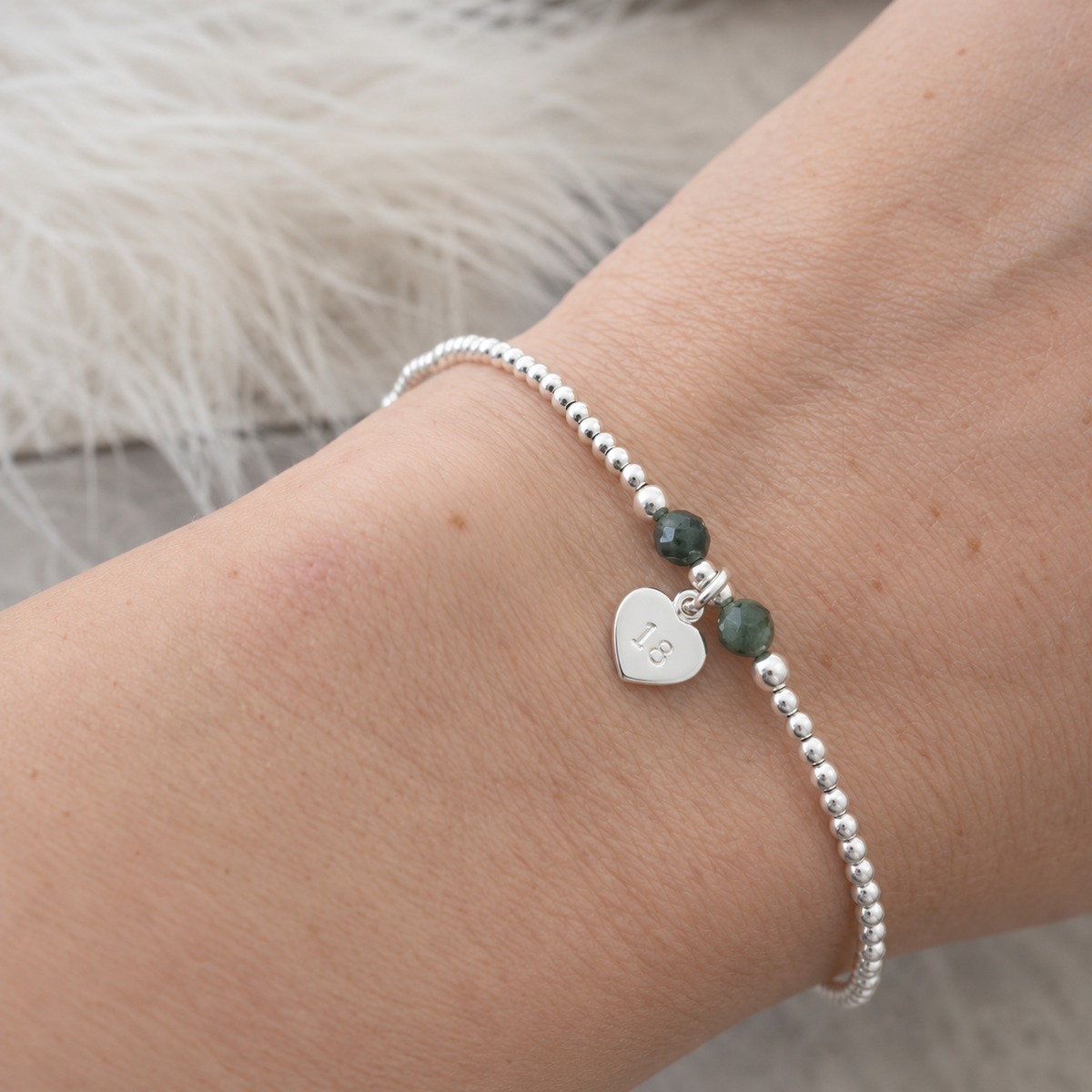 A silver bracelet with a heart-shaped charm and two green beads on a person's wrist.