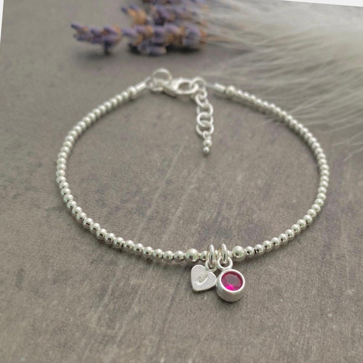 A silver-toned beaded bracelet with a charm and a small red gemstone is displayed on a gray surface.
