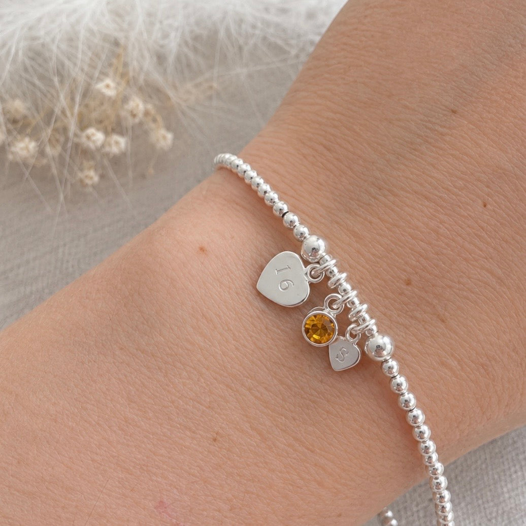 A silver bracelet with a heart-shaped charm and a small gemstone charm is worn on a person's wrist.