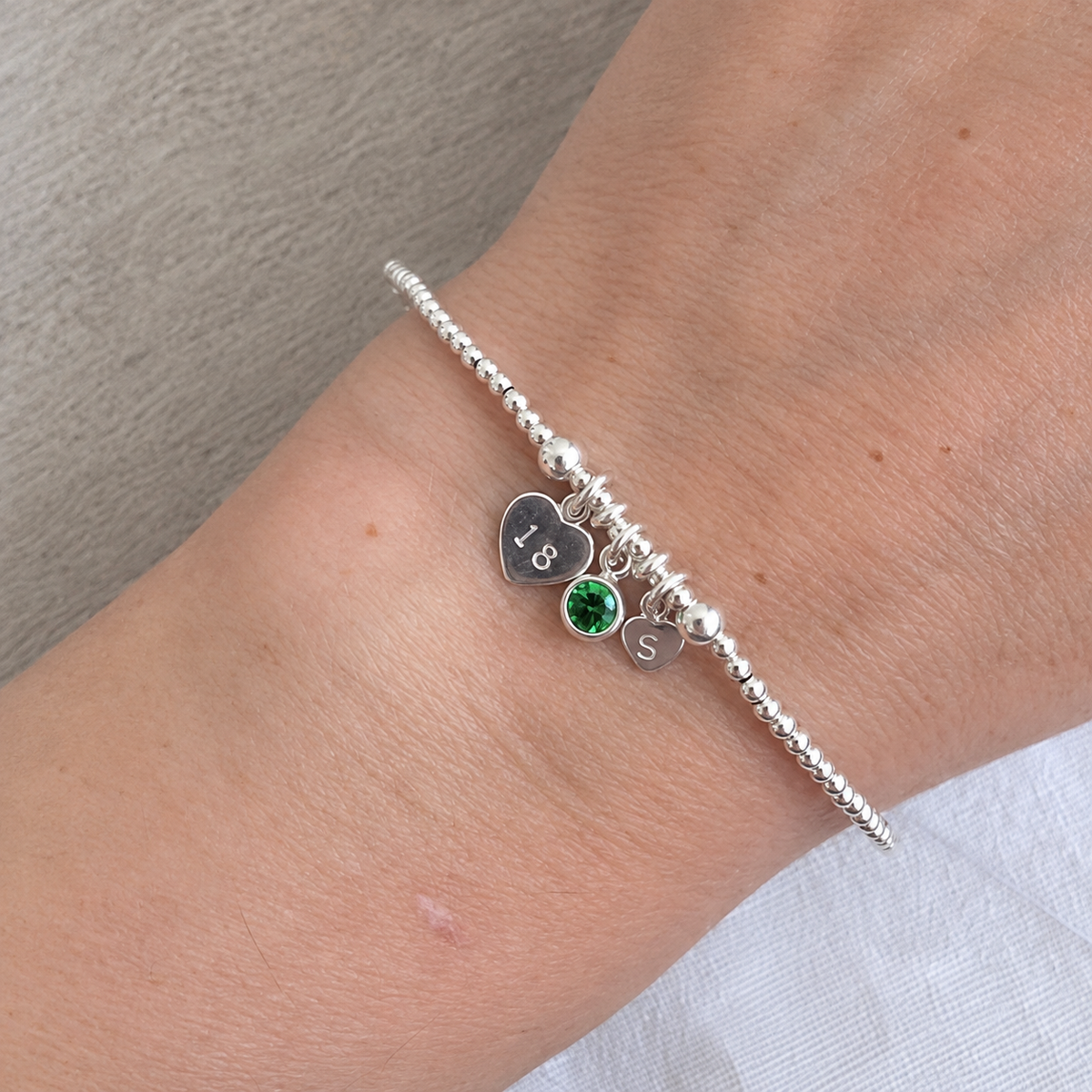 A silver bracelet with a heart charm and a green gemstone charm is worn on a person's wrist.