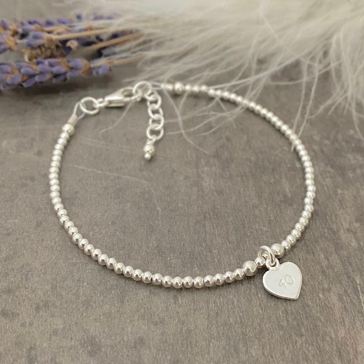 A silver bracelet with a heart-shaped charm and the number 10 engraved on it, placed on a gray surface with a feather-like object in the background.