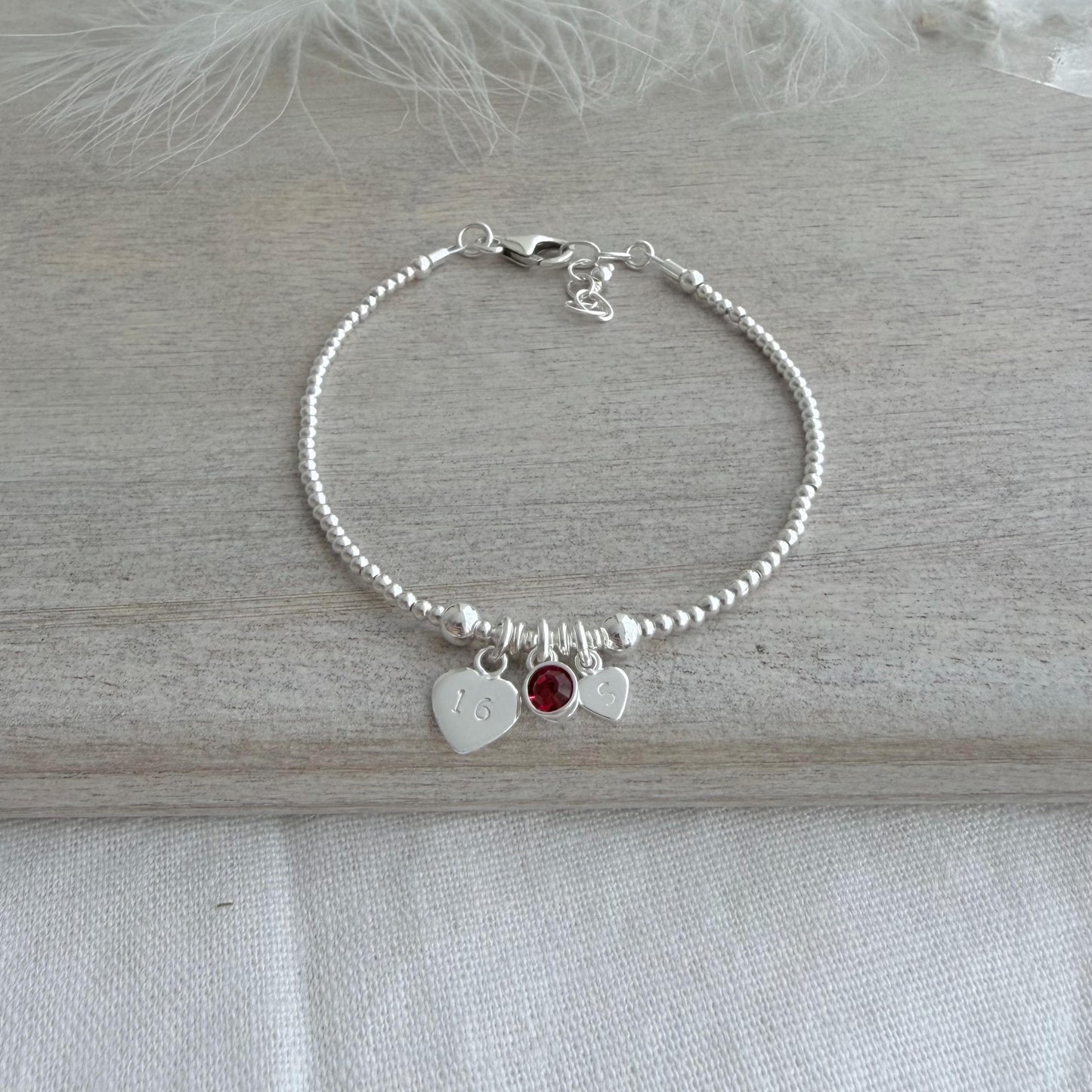 May include: A silver beaded bracelet featuring three charms. One heart charm is engraved with the number 16. A small red gemstone and a second heart charm are also attached. The bracelet rests on a light grey wooden surface.