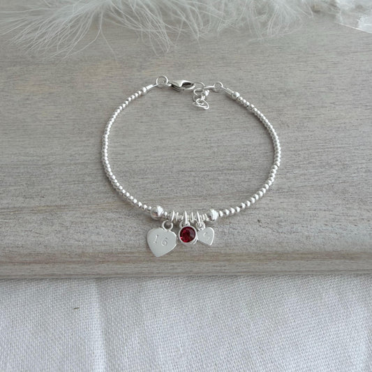 May include: A silver beaded bracelet featuring three charms. One heart charm is engraved with the number 16. A small red gemstone and a second heart charm are also attached. The bracelet rests on a light grey wooden surface.
