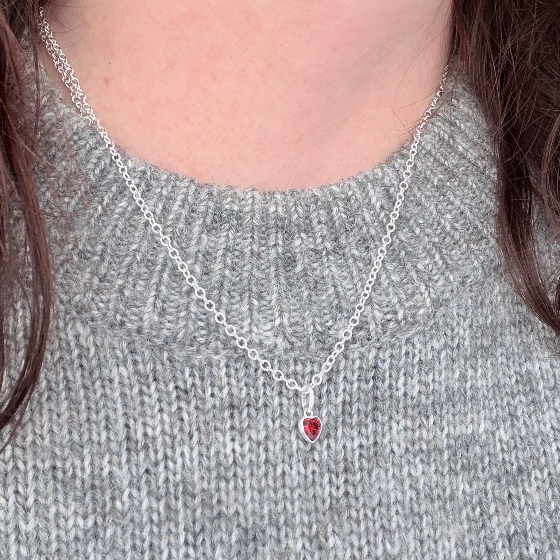 May include: A silver necklace featuring a small, heart-shaped red pendant. The pendant is a bright red and hangs from a delicate silver chain. The necklace is displayed against a grey jumper.