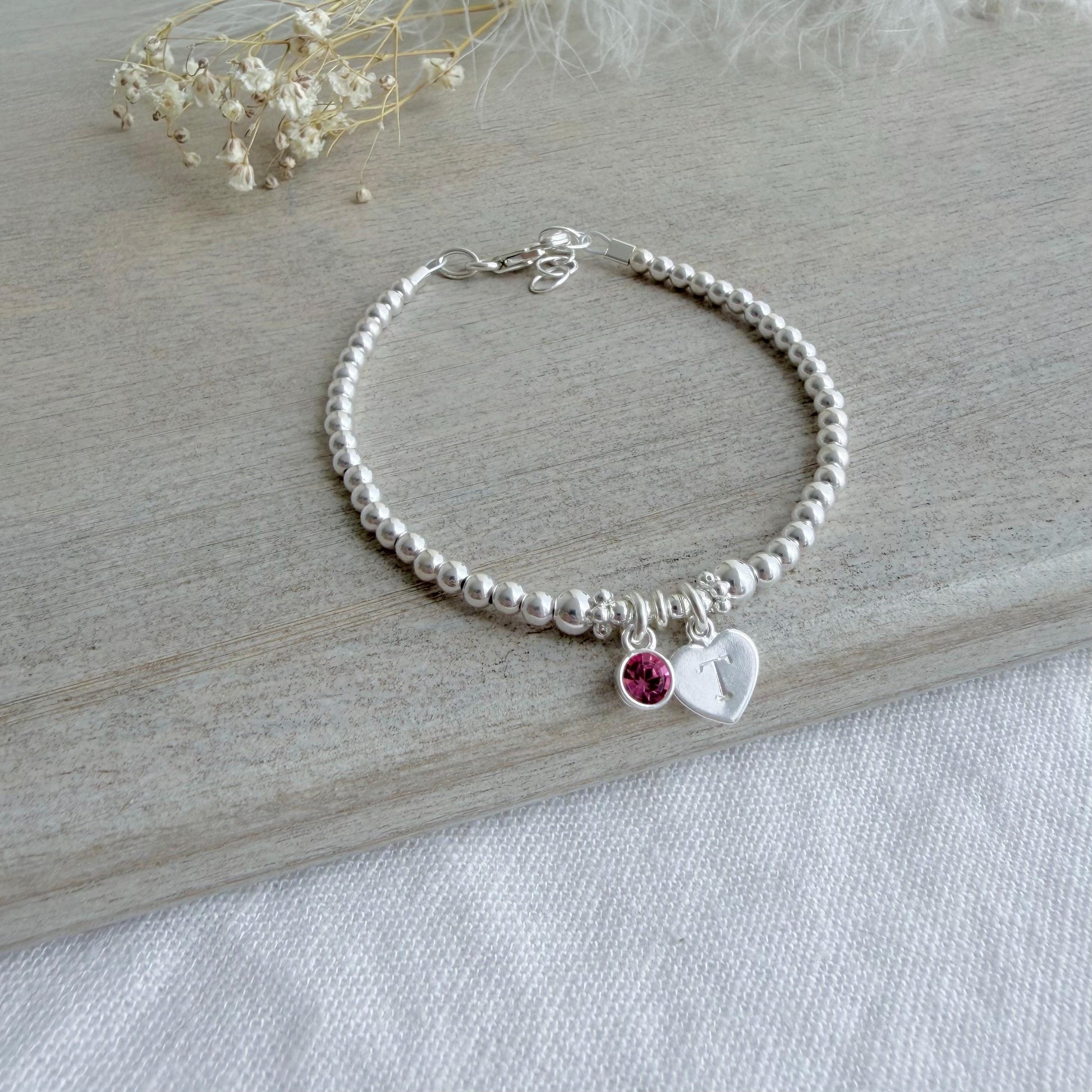May include: A silver beaded bracelet featuring a heart charm with the letter "T" and a small pink gemstone. The bracelet is displayed on a light wooden surface, with dried flowers in the background.
