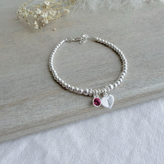 May include: A silver beaded bracelet featuring a heart charm with the letter "T" and a small pink gemstone. The bracelet is displayed on a light wooden surface, with dried flowers in the background.