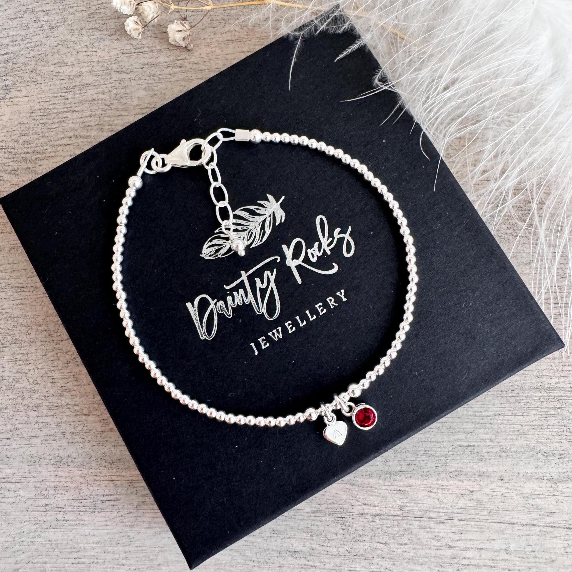 May include: A silver beaded bracelet with a heart charm and a red gemstone charm. The bracelet is presented on a black box with the text "Dainty Rocks Jewellery". The bracelet is made of small, round beads.