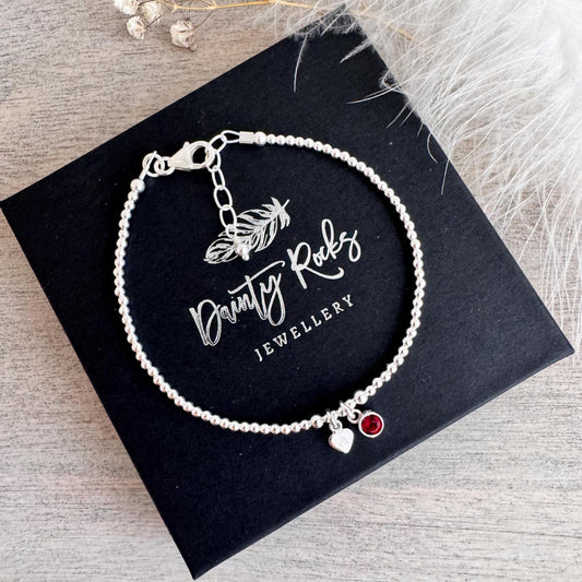 May include: A silver beaded bracelet with a heart charm and a red gemstone charm. The bracelet is presented on a black box with the text "Dainty Rocks Jewellery". The bracelet is made of small, round beads.