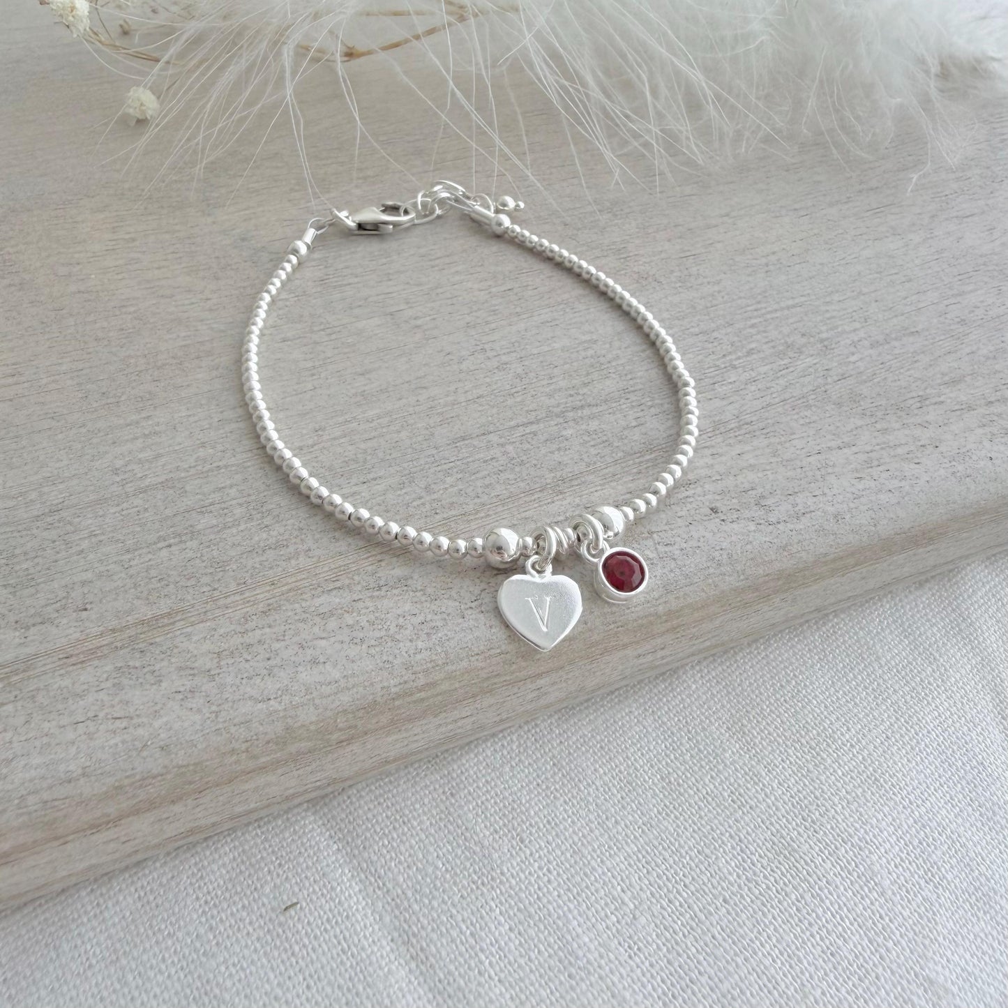May include: A silver bracelet featuring small beads, a heart-shaped charm with the letter "V", and a red gemstone. The bracelet is displayed on a light wooden surface.