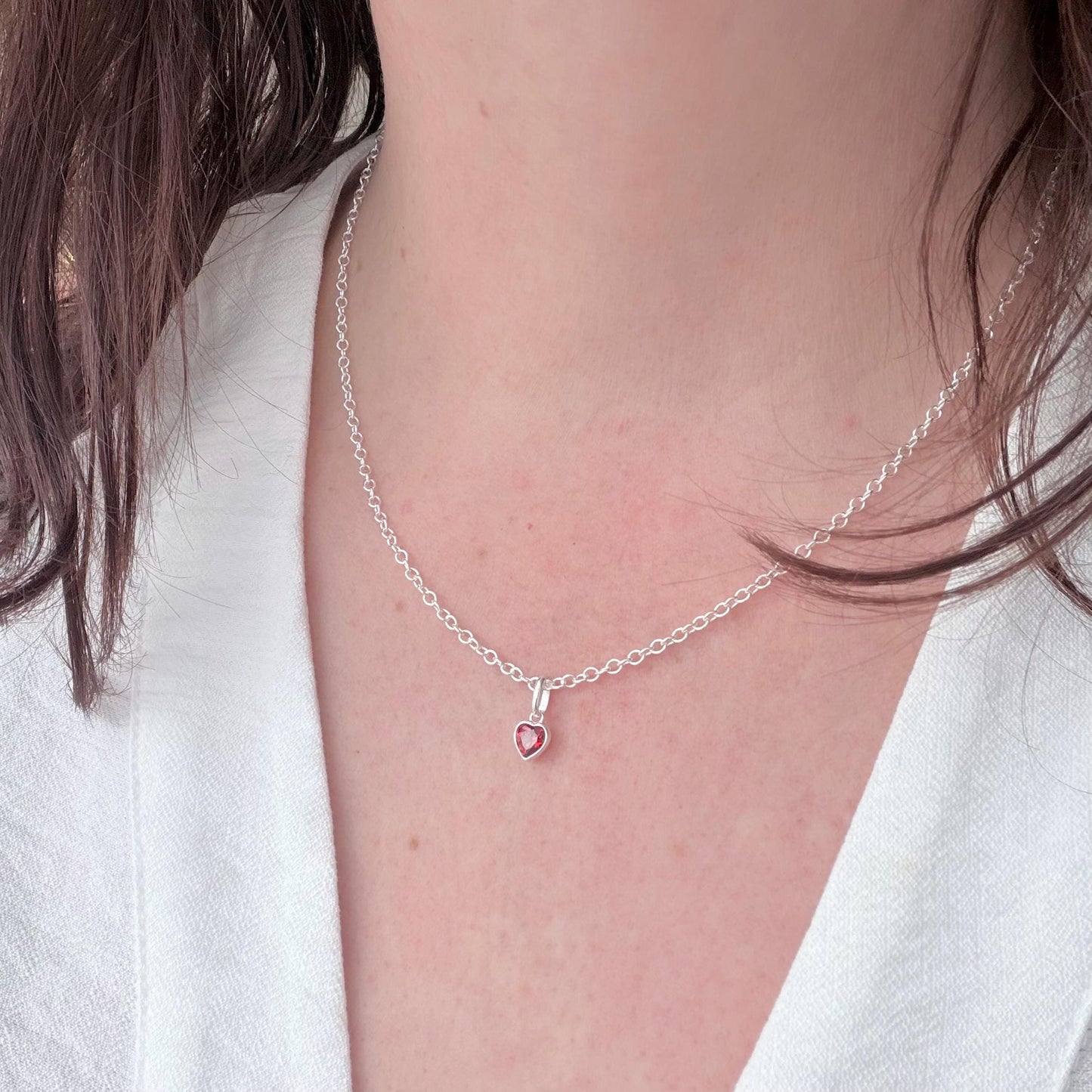 May include: A silver necklace with a small heart-shaped pendant, set with a red gemstone. The fine chain is visible against a white shirt. The pendant is a small, heart-shaped charm, creating a simple and elegant design.