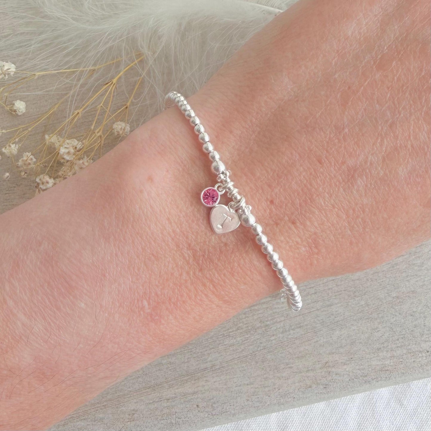 May include: A silver beaded bracelet featuring a heart charm and a pink gemstone. The heart charm is engraved with the letter "T". The bracelet is worn on a wrist.