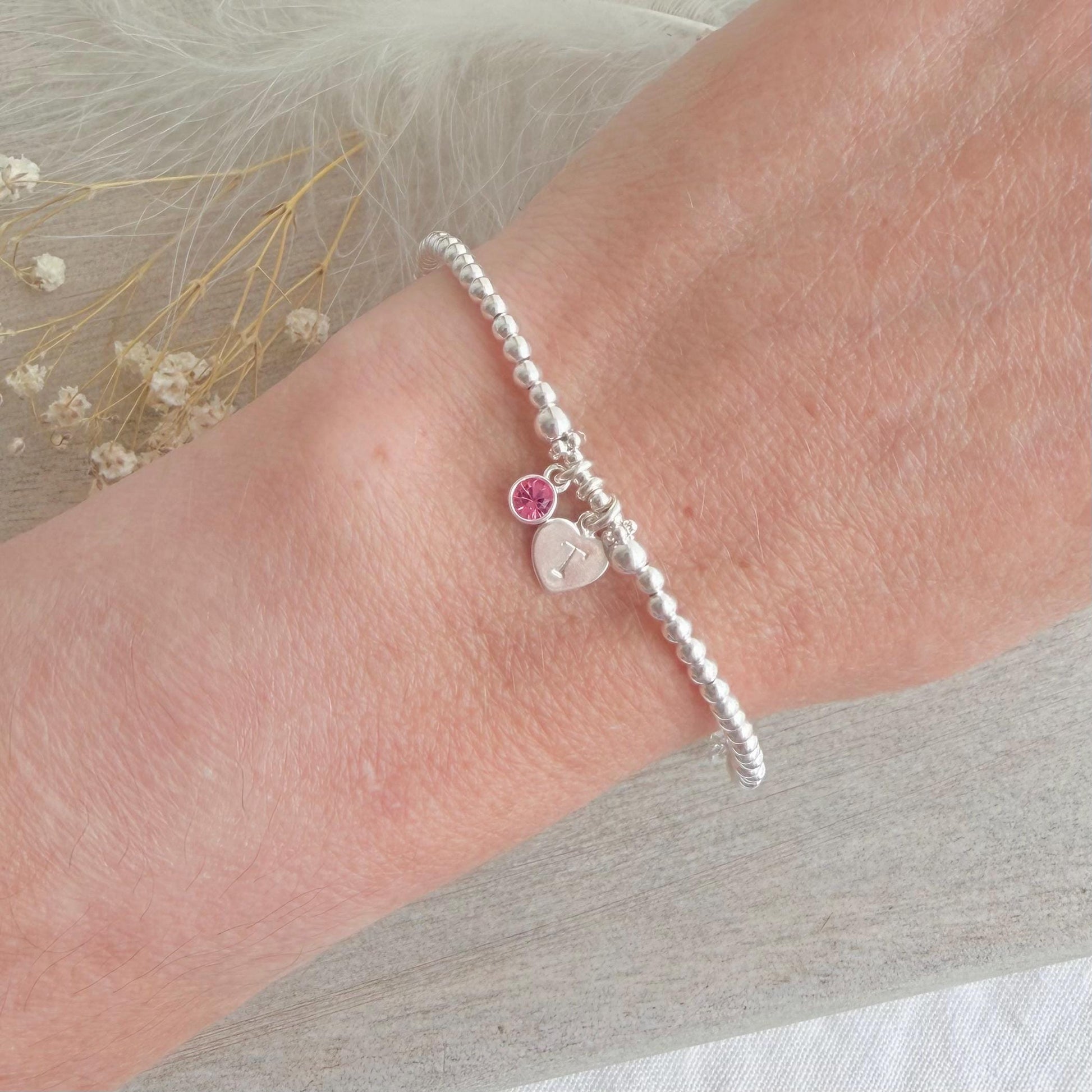 May include: A silver beaded bracelet featuring a heart charm and a pink gemstone. The heart charm is engraved with the letter "T". The bracelet is worn on a wrist.