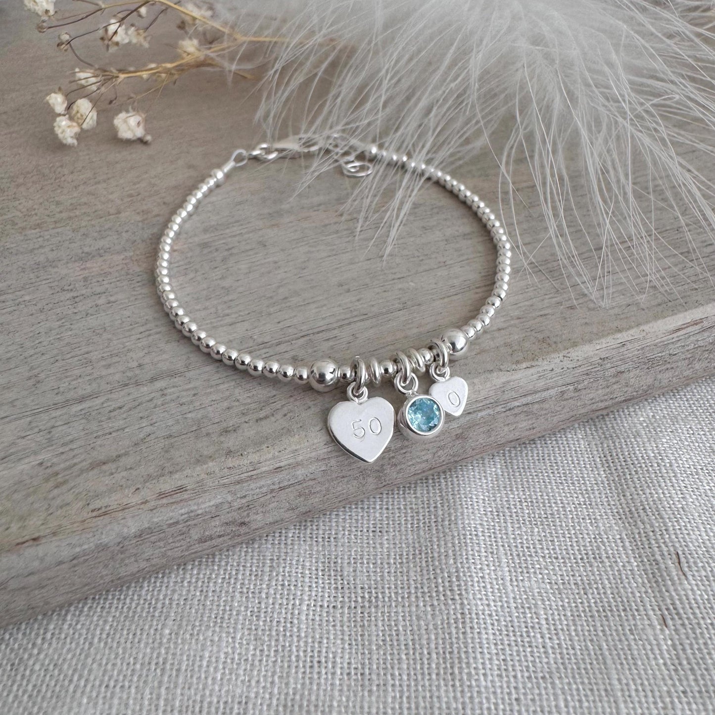 May include: A silver beaded bracelet featuring three charms. One heart charm is engraved with the number "50". A light blue gemstone and a small heart charm are also attached. The bracelet is presented on a wooden surface.
