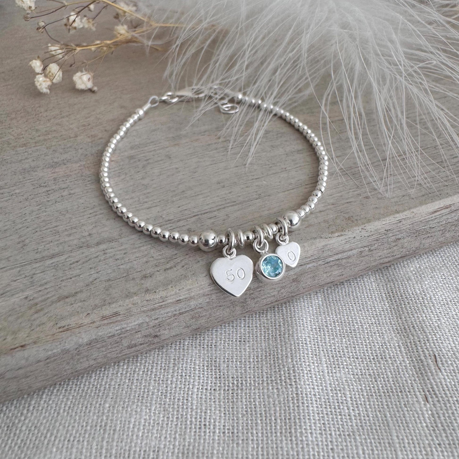 May include: A silver beaded bracelet featuring three charms. One heart charm is engraved with the number "50". A light blue gemstone and a small heart charm are also attached. The bracelet is presented on a wooden surface.