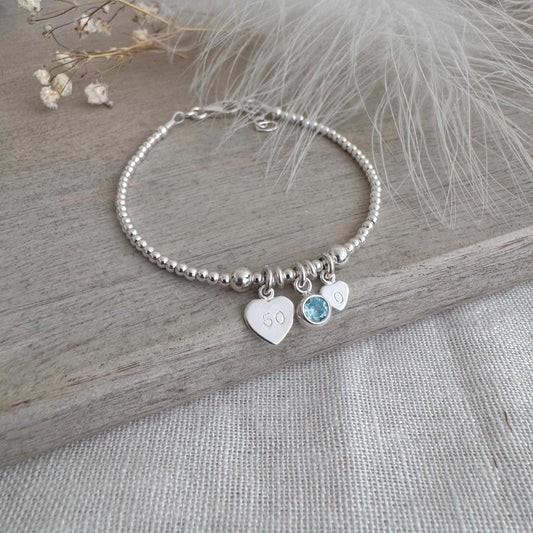 May include: A silver beaded bracelet featuring three charms. One heart charm is engraved with the number "50". A light blue gemstone and a small heart charm are also attached. The bracelet is presented on a wooden surface.