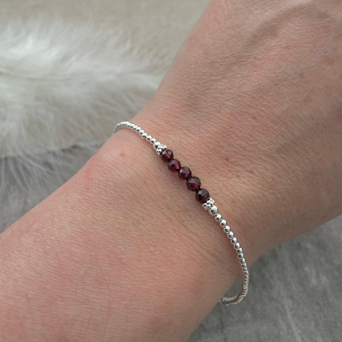 May include: A silver bracelet with small, round beads and a central cluster of five deep red garnet beads. The bracelet is displayed on a wrist, highlighting its understated elegance and subtle sparkle.