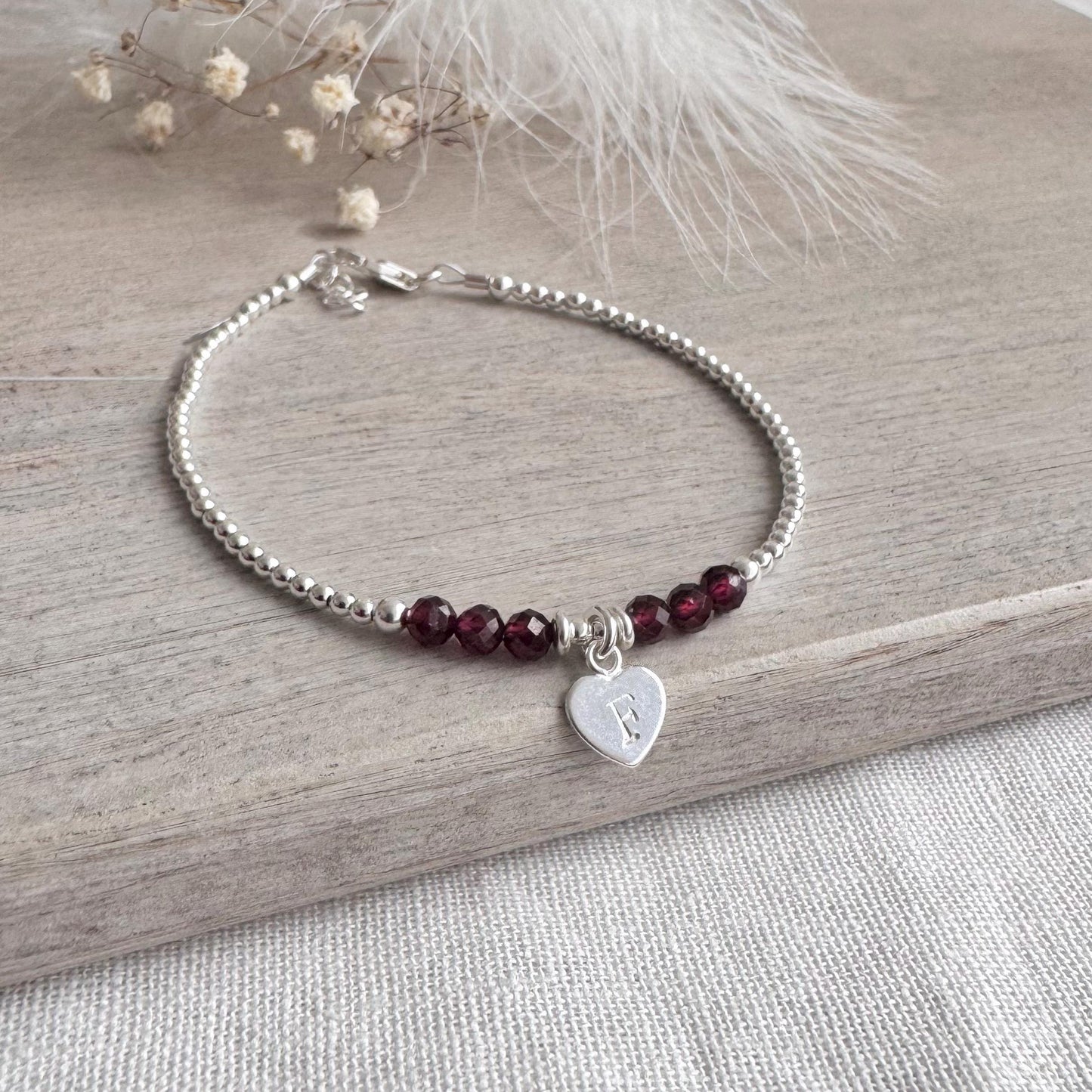May include: A silver bracelet with a heart-shaped charm bearing the letter "F". The bracelet is made of small silver beads and dark red beads. The bracelet is displayed on a wooden surface.
