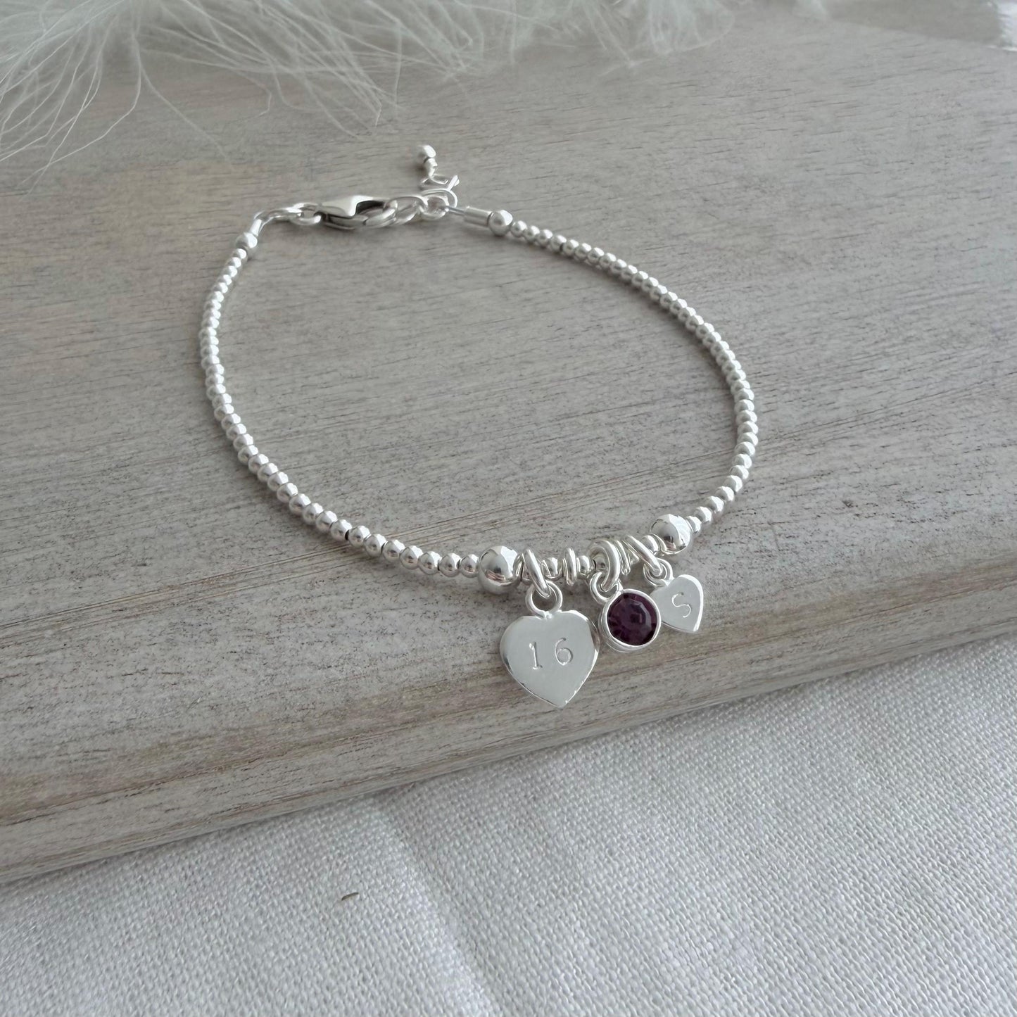 May include: A silver beaded bracelet featuring three charms. One heart charm is engraved with the number 16. A small purple gemstone and a heart charm with the letter "S" are also attached. The bracelet rests on a light grey wooden surface.
