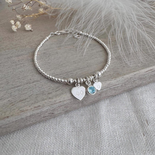 May include: A silver beaded bracelet featuring three charms. One heart charm is engraved with the number "50". A light blue gemstone and a small heart charm are also attached. The bracelet is presented on a wooden surface.